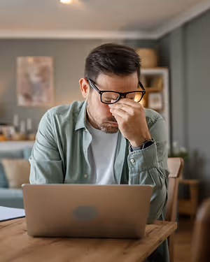A man wearing glasses is sitting on a chair in front of a table while covering his face with his hand, looking down at his laptop. He seems to be tired, and he is probably in his house. Behind him is a wooden table with a pillow and a shelf with a picture frame and some stuff on it. There is also a couch with pillows and a lamp on the left.