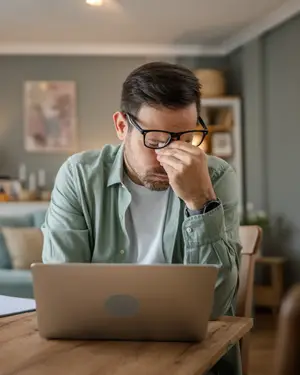 A man wearing glasses is sitting on a chair in front of a table while covering his face with his hand, looking down at his laptop. He seems to be tired, and he is probably in his house. Behind him is a wooden table with a pillow and a shelf with a picture frame and some stuff on it. There is also a couch with pillows and a lamp on the left.