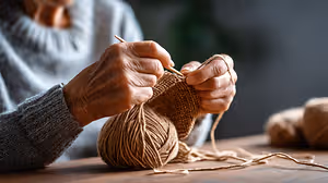 An elderly woman with gray hair is knitting with yarn on a wooden table, a blurry background of a potted plant is behind her.
