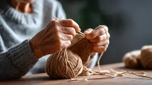 An elderly woman with gray hair is knitting with yarn on a wooden table, a blurry background of a potted plant is behind her.