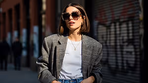 A woman with short hair wearing sunglasses, a necklace, and a plaid blazer stands on a sidewalk with buildings and graffiti in the background.