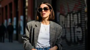 A woman with short hair wearing sunglasses, a necklace, and a plaid blazer stands on a sidewalk with buildings and graffiti in the background.