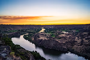 A wide landscape view of a river flowing through a canyon with a sunset in the background.