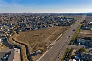 A wide aerial view of a residential neighborhood with a large open field in the middle, bordered by houses, and a road on the right side. There are vehicles on the road and some houses on the left side.