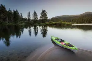A green kayak is parked on the shore of a lake with reflections of trees and mountains in the water.