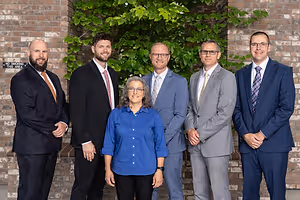 Five people dressed in suits and glasses standing in front of a brick wall with a plant behind them