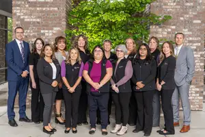 A group of people standing in front of a brick wall with a tree behind them, all dressed in business attire and smiling for a photograph.