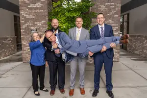 Five people in formal attire posing for a photo outside a building with a brick wall and a tree in the background.