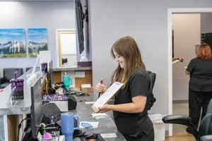 A woman standing in front of a desk with a clipboard, smiling and holding a pen, with other people and items in the background.