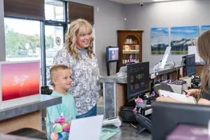 Two adults and a child are standing at a reception desk in a salon. One of the adults is smiling and appears to be conversing with the child. The desk is cluttered with various items like a monitor, a handbag, a glass bowl, a book, a pen, and a basket. A surveillance camera is mounted on the wall. The reception area is well-lit and has a glass door and window with blinds.