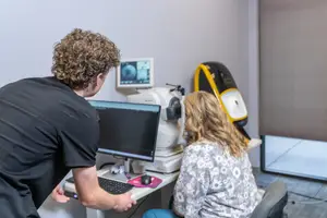 A man and woman in an eye clinic, with the man looking at a monitor while the woman sits in front of an autorefractor and optical biometer machine.