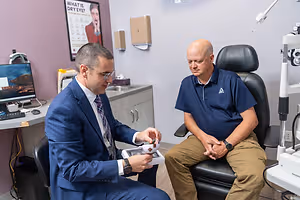 Two men are sitting in an eye exam room. One man is holding a device while the other looks on.