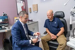 Two men are sitting in an eye exam room. One man is holding a device while the other looks on.