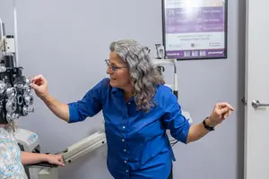 An older woman in a blue shirt is adjusting a child's eye exam equipment while smiling