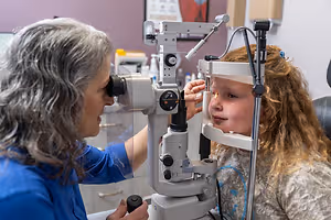 An eye doctor examining a young girl's eyes with a phoropter in an exam room.