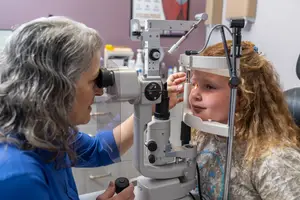 An eye doctor examining a young girl's eyes with a phoropter in an exam room.