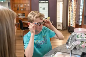 A young boy in a blue shirt with a geometric design is trying on glasses at an optical shop, with a woman looking at him and a mirror and boxes in the background.