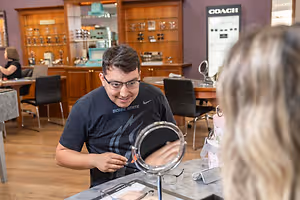 A man in glasses is helping a woman try on glasses in an eyeglass store