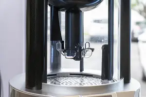 A pair of eyeglasses is inside a black and silver machine with a glass window, probably a lens polishing machine, and a blurry background of a car and trees.