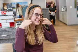 A girl with glasses and a headband smiles and adjusts her earpiece while sitting in a room with wooden floors and a cabinet with books