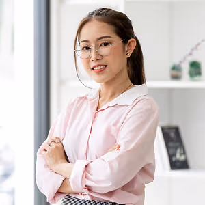 Woman wearing eyeglasses and a pink shirt with a white collar standing in an office with a smile on her face.