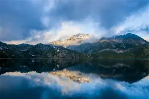 A serene landscape with a lake reflecting the mountains and sky with clouds.