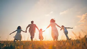 A family of four, including two adults and two children, is joyfully running together in a field as the sun sets, casting a warm glow over them.