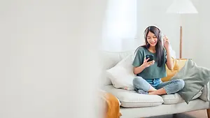A woman sitting on a couch wearing headphones and holding a cell phone with a smile on her face