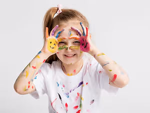 A young girl is smiling and posing with her hands covered in paint, probably for a photo. She has a white t-shirt on, and her hands are covered in different colors of paint. She has a ponytail hairstyle, and her face is slightly dirty with paint. She is probably standing in a room, and the wall behind her is white.