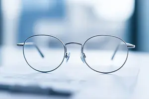 A pair of glasses with clear lenses and silver frames is placed on a white surface, possibly a desk, with a blurry background of a blue wall and a window.