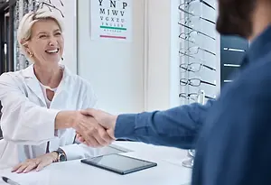 An adult female sitting at a desk shakes hands with an adult male in a lab coat