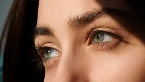 A woman with long black hair and green eyes is looking to the left, with her face partially illuminated by the sunlight. She has long, thick eyelashes and her eyes are slightly closed.