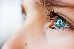 Close-up of a woman's face with a blurry background showing her eye with green and blue irises and long lashes.