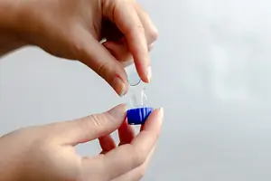 A person is holding a small transparent plastic container with a blue lid, possibly demonstrating how to insert a contact lens into the container.