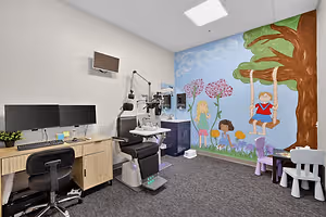 A room with a computer desk, a chair, a wall mural, a sink, and a television mounted on the wall, with a large tree and flowers painted on the wall