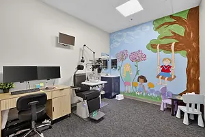 A room with a computer desk, a chair, a wall mural, a sink, and a television mounted on the wall, with a large tree and flowers painted on the wall