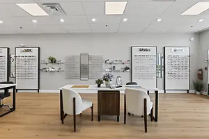 An image of a desk with chairs and plants inside a shop, with a mirror and shelves on the wall with eyeglasses displayed, along with a fire extinguisher and a white wall with ceiling lights