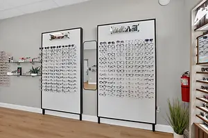 A white room with a display of glasses on a wall and a mirror on the side and a shelf with a potted plant on it.