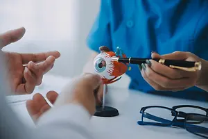 A person with a blue shirt is pointing at a toy eyeball with a pen in their hand on a white table.