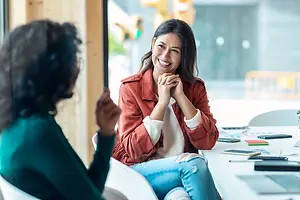 A woman smiles and laughs while sitting in a chair in an office with a desk and another person sitting across from her