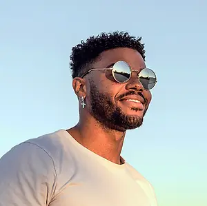 A smiling black man in sunglasses and a white shirt poses for a picture.