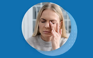 A woman with blonde hair is covering her face with her hands and appears to be in pain. She is probably sitting inside a house. The blue background has a blue circle with a white dot pattern in the middle. The woman is probably suffering from a headache.