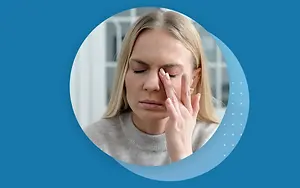 A woman with blonde hair is covering her face with her hands and appears to be in pain. She is probably sitting inside a house. The blue background has a blue circle with a white dot pattern in the middle. The woman is probably suffering from a headache.