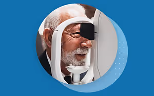 An elderly man wearing a black eye patch is using an eye examination machine while smiling at the camera