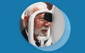 An elderly man wearing a black eye patch is using an eye examination machine while smiling at the camera