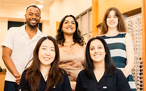 Five people are posing for a picture in an office with a wall of eyeglasses behind them
