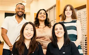 Five people are posing for a picture in an office with a wall of eyeglasses behind them