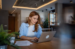 A woman sitting at a desk, working on a laptop, with a cup, a potted plant, and papers in front of her