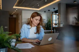 A woman sitting at a desk, working on a laptop, with a cup, a potted plant, and papers in front of her