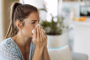 A woman is sitting on a couch and blowing her nose with a tissue while looking down.